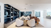 Located at 250 Plaza Circle in Mundelein, IL, this contemporary community living room offers a generously sized, single-floor open layout thoughtfully designed for both relaxation and entertainment. The space is defined by a clean, modern aesthetic, featuring crisp white ceilings, neutral-toned flooring, and bold accent walls in deep teal and warm terracotta that add visual depth and character throughout.

The main lounge area is anchored by a large built-in shelving unit finished in slate blue-gray, housing a flat-screen TV, decorative objects, and curated accessories. Comfortable seating arrangements — including a tufted brown sofa, plush white swivel chairs, and gold-accented side tables — are artfully grouped atop a patterned area rug, creating an inviting social hub. A sculptural pendant light adds an artistic focal point overhead.

An adjacent game zone features a ping-pong table, high-top seating, an arcade machine, a tabletop game station, and a classic popcorn cart, lending the space a playful, recreational energy. Framed artwork punctuates the teal accent walls throughout. Large glazed entry doors and windows allow natural light to flow in, enhancing the bright, airy ambiance. The overall design balances sophisticated style with approachable comfort, making this an ideal communal gathering space.
