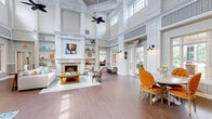 Located at 101 Sabal Palm Ln in Chesapeake, Virginia, this single-story clubhouse facility at MAA Adalay Bay offers a thoughtfully designed community space spanning a generous footprint. The centerpiece of the facility is a dramatic main living area boasting soaring double-height ceilings, intricate wainscoting, and rich hardwood flooring. Towering clerestory windows flood the space with an abundance of natural light, while a stately fireplace framed by built-in shelving serves as an elegant focal point. Ceiling fans complement the airy atmosphere throughout. A dining area with vibrant accent chairs and a round table sits adjacent to the main lounge, creating a seamless, open flow. A secondary living room features warm, intimate décor with bold terracotta walls, colorful artwork, and cozy seating arrangements. The hallway connects all areas efficiently, while a dedicated office space, finished with patterned carpeting and classic furnishings, provides a quiet professional environment. The fitness center is exceptionally well-equipped, featuring Matrix cable machines, free weights, treadmills, ellipticals, and stationary bikes beneath a vaulted ceiling with large windows overlooking the outdoors. Mirrored walls and rubber flooring complete this modern exercise space. Together, these amenity spaces deliver a welcoming, upscale community experience.