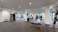 Located at 7510 N Oak Trafficway in Gladstone, Missouri, this fitness center offers a comprehensive, single-floor workout facility thoughtfully divided into two distinct training areas. The main workout floor is generously proportioned, featuring rubber sport flooring, recessed ceiling lighting, and structural columns that define the open layout without restricting movement. Floor-to-ceiling black-framed windows line one wall, flooding the cardio zone with natural light and providing refreshing views of the surrounding greenery — an inviting backdrop for treadmills, elliptical machines, and rowing equipment. Full-length mirrors along another wall visually expand the space and support proper form during strength training. A dedicated free weights area includes a squat rack with Olympic barbells and a dumbbell station, catering to a range of strength training needs. A separate, enclosed room serves as a functional training studio, outfitted with a heavy punching bag, medicine balls, kettlebells, TRX suspension trainers, and battle rope equipment — ideal for high-intensity and cross-training workouts. This room features carpet tile flooring and bright overhead lighting. Together, the two zones provide a well-rounded fitness environment suitable for cardio, strength, and functional training disciplines.