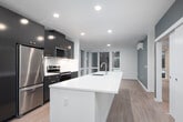 This contemporary single-floor unit offers a thoughtfully designed open-concept layout that seamlessly integrates the kitchen, dining, and living areas. The kitchen is a standout feature, anchored by a generous white quartz waterfall island with an undermount stainless steel sink. Dark espresso cabinetry with warm gold hardware provides striking contrast against the white tile backsplash and light quartz countertops. Stainless steel appliances — including a full-size refrigerator, electric range, and over-the-range microwave — complete the well-appointed kitchen. Recessed lighting throughout ensures consistent, modern illumination. Light wood-tone flooring extends across the entire unit, contributing to a warm and cohesive aesthetic. The open living area features a slate-blue accent wall and a wall-mounted ductless mini-split system for climate control, with large windows offering views of the surrounding urban environment. A sliding door leads to additional rooms, including a spacious bathroom finished with a dark vanity, dual undermount sinks, white quartz countertops, a full bathtub and shower, and a stacked in-unit washer and dryer. The overall ambiance is clean, contemporary, and functional, designed to maximize comfort and utility.