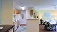 This single-floor residence offers a practical and welcoming layout centered around an open-concept design that seamlessly connects the kitchen and living areas. The kitchen is well-appointed with light wood cabinetry, white appliances including a refrigerator and range, and a peninsula countertop that serves as both a workspace and a casual divider between the kitchen and the adjoining living space. A skylight-style ceiling fixture brightens the kitchen, complementing the natural flow of light throughout. The flooring throughout is a warm wood-style laminate, adding cohesion and a grounded aesthetic to the space. Soft green-tinted walls in the living area contribute a calm, neutral ambiance. An electrical panel is conveniently recessed into the wall, maintaining a clean and organized appearance. The living area, visible through the pass-through opening, is spacious enough to accommodate comfortable furnishings and receives additional light from nearby fixtures. The overall layout is efficient and thoughtfully arranged, making excellent use of the available footprint while maintaining an open, airy feel throughout the main living spaces.