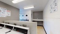 This single-floor shared laundry facility is designed for practical, high-capacity use. The room is outfitted with a row of Speed Queen commercial front-load washing machines along one wall, paired with stacked commercial dryers on the opposite side, offering ample capacity for multiple users simultaneously. The layout is efficiently organized, allowing clear circulation between the machines.

The interior features neutral gray and white walls with light-toned flooring, creating a clean, functional environment. Fluorescent strip lighting runs across the ceiling, ensuring the space is well-lit throughout. A window along one wall admits natural light and provides a view of the surrounding outdoor area, adding a degree of openness to the otherwise utilitarian room.

A built-in white folding shelf is positioned along one wall, providing a dedicated surface for sorting and folding laundry. Instructional signage for both washing and drying is posted prominently on the walls for user reference. A fire extinguisher is mounted near the entrance, and a waste bin is conveniently placed near the folding counter. The entrance door features a small window, and CSC service information is displayed for maintenance contact purposes, reflecting a well-managed, community-oriented facility.
