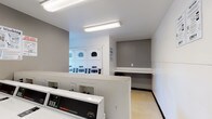 This single-floor shared laundry facility is designed for practical, high-capacity use. The room is outfitted with a row of Speed Queen commercial front-load washing machines along one wall, paired with stacked commercial dryers on the opposite side, offering ample capacity for multiple users simultaneously. The layout is efficiently organized, allowing clear circulation between the machines.

The interior features neutral gray and white walls with light-toned flooring, creating a clean, functional environment. Fluorescent strip lighting runs across the ceiling, ensuring the space is well-lit throughout. A window along one wall admits natural light and provides a view of the surrounding outdoor area, adding a degree of openness to the otherwise utilitarian room.

A built-in white folding shelf is positioned along one wall, providing a dedicated surface for sorting and folding laundry. Instructional signage for both washing and drying is posted prominently on the walls for user reference. A fire extinguisher is mounted near the entrance, and a waste bin is conveniently placed near the folding counter. The entrance door features a small window, and CSC service information is displayed for maintenance contact purposes, reflecting a well-managed, community-oriented facility.
