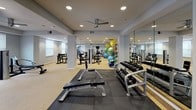This fitness center is a comprehensive, single-level facility designed to accommodate a wide range of workout routines. The space is organized into distinct functional zones, allowing users to move seamlessly between cardio, strength training, and open-floor exercise areas. The cardio section houses multiple treadmills, elliptical trainers, and a stationary bike, positioned to face a wall-mounted television for an engaging workout experience. A dedicated free-weight area features a tiered dumbbell rack alongside a flat bench and protective rubber flooring, complemented by a large wall mirror that enhances the sense of depth and allows users to monitor their form. The strength training section includes a cable machine and additional benches, while a separate open area accommodates stability balls and functional training accessories. Neutral gray walls, recessed ceiling lighting, and a ceiling fan contribute to a clean, comfortable atmosphere throughout. Carpeted zones transition smoothly into rubber-matted workout areas, providing appropriate surfaces for varied activities. French doors at the rear of the space open to an outdoor area with views of a pool, introducing natural light and convenient access to additional amenities. A water cooler station is also conveniently located within the facility.