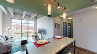 This contemporary urban unit showcases a thoughtfully designed open-concept layout with distinct industrial-modern character throughout. The spacious living area immediately impresses with its soaring exposed steel beam ceilings, complete with visible ductwork that adds an authentic loft-like aesthetic. Floor-to-ceiling windows flood the space with natural light and frame dynamic views of the surrounding cityscape, creating a vibrant connection to the urban environment outside.

The well-appointed kitchen is a standout feature, boasting light maple cabinetry extending to the ceiling, stainless steel appliances including a full-size refrigerator, stove, microwave, and dishwasher, complemented by generous counter space and a bold green-painted ceiling with track lighting. A built-in dining island with seating bridges the kitchen and living areas seamlessly.

Two private bedrooms each offer natural light through bright windows overlooking the city, with comfortable carpet or hardwood-style flooring. The shared bathroom features a dual-sink vanity with light wood cabinetry, a wide mirror, and clean white countertops, providing ample space for multiple occupants. Hardwood-style flooring runs throughout the main living areas, tying together a cohesive, modern aesthetic ideal for urban living.