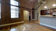 This distinctive single-floor loft showcases an impressive blend of industrial heritage and contemporary design. The open living area is anchored by original exposed brick walls that rise to soaring ceilings, lending the space a raw, authentic character. A dramatic brick archway serves as a defining architectural feature, seamlessly connecting the main living area to an adjoining room and adding a sense of grandeur to the layout. Polished hardwood floors run throughout, reflecting natural light and complementing the warm tones of the brick. Large multi-pane windows flood the space with abundant natural light, enhancing the open, airy atmosphere. Exposed metal ductwork traces the ceiling, reinforcing the space's industrial aesthetic. On one side, rich dark-wood cabinetry and a granite-topped counter mark the kitchen area, offering a refined contrast to the surrounding raw elements. Additional doorways lead to separate rooms, providing functional separation within the otherwise open floor plan. The overall ambiance is one of spacious, loft-style living where historic architectural details and modern finishes coexist harmoniously, making for a visually dynamic and highly characterful space.