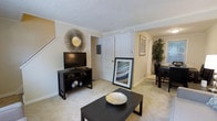 This two-story residence offers a well-organized layout designed for comfortable everyday living. The main level is anchored by a carpeted living room that sets a warm, inviting tone throughout the space. Crown molding adds a refined architectural detail along the ceiling, complementing the textured ceiling finish above. A staircase positioned near the living area provides convenient access to the upper floor.

The living room flows naturally into an adjacent dining area, visible through an open corridor that enhances the sense of connectedness between the spaces. The dining area accommodates a full table and chairs, with a decorative indoor plant adding a touch of greenery. Framed artwork and wall décor contribute to the overall aesthetic.

A white paneled door leads from the main living area to additional spaces, while neutral-toned walls and light carpeting throughout create a cohesive, understated palette. The layout appears designed to maximize usability across both floors, with the main level serving as the primary living and dining hub. Overall, the space presents a clean, functional interior with classic residential detailing.