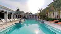 This outdoor amenity space centers around a generously sized swimming pool with vivid turquoise water, creating a resort-like atmosphere reminiscent of a luxury retreat. The pool is flanked by decorative mosaic-tiled pillars and smooth stone spheres, adding sculptural elegance to the surroundings. A wide pool deck provides ample space for lounging, lined with chaise lounge chairs positioned to take advantage of the open sky and abundant natural light.

The backdrop features a well-appointed single-story clubhouse with a curved architectural element and large windows that allow natural light to flow through its interior. Orange market umbrellas dot the deck area, offering shaded seating for residents or guests. The structure's clean, modern facade is complemented by mature tropical palm trees that frame the space and contribute to its warm, coastal ambiance.

Overall, the space exudes a polished, tropical aesthetic with thoughtful landscaping, high-quality finishes, and an open layout designed to encourage outdoor relaxation and social gathering in a sophisticated setting.