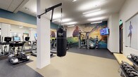 This fitness center is designed to support a wide range of workout styles within a single, open-plan floor. The space is anchored by a striking large-scale athletic mural that spans the rear wall, depicting dynamic sports imagery in vivid color and creating an energizing visual backdrop that sets the tone for the entire room.

The layout is thoughtfully organized to accommodate both cardio and strength training. A row of cardio machines, including treadmills and stationary bikes, lines one side of the space, each equipped with individual screens for entertainment or workout tracking. On the opposite end, a multi-function cable strength station provides versatile resistance training options, complemented by a set of medicine balls and a stability ball for functional fitness exercises.

A wall-mounted pull-up bar supports an Everlast heavy punching bag, adding a dedicated boxing and combat training element to the space. Recessed and panel lighting throughout ensures the room is consistently well-lit. The flooring transitions between durable vinyl and dark rubberized mat sections, delineating workout zones while providing appropriate support and traction. A wall-mounted TV adds a further entertainment option, rounding out this comprehensive fitness facility.