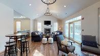 This single-floor community space is thoughtfully designed to encourage socializing and comfort across several distinct zones. The centerpiece is a spacious main living area with soaring vaulted ceilings that lend an open, airy feel. A statement chandelier hangs above two tufted navy sofas arranged around a dark wood coffee table, with a tile-surround gas fireplace and wall-mounted television serving as the focal point. Large windows and a set of glass French doors flood the room with natural light and offer views of the outdoors. Rich hardwood-style flooring flows throughout, anchoring the warm, neutral color palette. Adjacent to the main seating area is a cozy secondary lounge featuring coordinating upholstered chairs arranged around a round side table — ideal for more intimate conversation. The kitchen is well-appointed with warm wood cabinetry, granite countertops, and black appliances including a range, microwave, and full-size refrigerator. A peninsula with bar stool seating connects the kitchen to the common area, making it convenient for casual dining or entertaining. A casual dining nook with high-top tables and stools provides additional flexible seating. Together, these spaces create a functional and inviting environment suited for a variety of community gatherings and events.