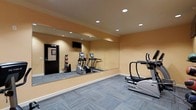 This fitness center is a purpose-built exercise space designed to support a well-rounded cardio and strength training routine. The room features warm, neutral-toned walls that create an inviting and energizing atmosphere, complemented by patterned grey carpeting that adds a modern, polished look underfoot. A large wall-to-wall mirror spans one side of the room, visually expanding the space and allowing users to monitor their form during workouts. Recessed ceiling lights provide bright, even illumination throughout, ensuring a well-lit environment at all hours. The facility is equipped with multiple cardio machines, including treadmills and an elliptical trainer, offering a variety of options for cardiovascular exercise. A wall-mounted flat-screen television is positioned for easy viewing during workouts. Additional fitness accessories, including resistance equipment, are stored neatly near the entrance. A dedicated entry door provides easy access to the facility. The overall layout is thoughtfully organized to maximize usable floor space, making the room feel open and functional. This fitness center is well-suited for daily exercise and promotes an active, healthy lifestyle in a clean and well-maintained environment.