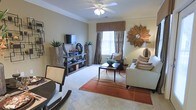 This single-floor residence showcases a thoughtfully designed living area that balances comfort and style. The living room is defined by elegant crown molding along the ceiling, adding a refined architectural touch, and a ceiling fan that enhances both function and form. Neutral-toned walls and plush carpeting create a warm, inviting atmosphere throughout the space. A large window dressed with layered curtains and a valance in earthy tones allows natural light to fill the room while maintaining privacy. The layout accommodates both a seating area and a dining space, demonstrating versatile use of the available floor plan. A media console with open shelving provides practical storage, complemented by woven basket accents and potted greenery that introduce natural texture. The seating arrangement centers around a dark-framed coffee table and is anchored by a rich terracotta-toned area rug that adds depth and warmth. Decorative elements, including a sculptural sunburst mirror and coordinated accent pillows, lend the space a polished, cohesive aesthetic. Overall, the space presents a comfortable and well-finished interior with attention to both design detail and livability.