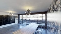 This single-floor space showcases a compelling blend of industrial and modern design. The most prominent feature is the expansive floor-to-ceiling steel-framed glass wall, which floods the interior with abundant natural light and provides unobstructed views of the surrounding landscaped exterior. The polished concrete flooring anchors the space with a clean, utilitarian aesthetic that complements the industrial character throughout.

A statement sputnik chandelier with exposed Edison-style bulbs serves as a focal point overhead, adding a retro-modern touch that contrasts effectively with the raw architectural elements. A long, light wood communal table paired with modern transparent chairs creates a functional workspace or meeting area at the heart of the room.

The space also features dark-framed wall-mounted organizational panels and signage, contributing to a professional, branded atmosphere. Textured stone or brick accent walls add depth and visual interest. The overall ambiance is open, contemporary, and purposeful — well-suited for a creative office, co-working environment, or collaborative workspace.