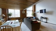 This two-floor space showcases a distinctive industrial aesthetic, blending raw architectural elements with warm, inviting finishes. The living area is immediately striking, with a rich wood-paneled ceiling that adds warmth and texture, complemented by exposed brick walls that lend a robust, urban character to the interior. Large windows on multiple walls flood the space with natural light, offering views of the surrounding trees and neighborhood outside.

The open layout of the main living area comfortably accommodates a generous sectional sofa arrangement, creating an inviting lounging space centered around a low coffee table. A dining area sits adjacent, furnished with a light wood table and chairs, making it well-suited for everyday meals or entertaining. The neutral tones of the carpet and walls are offset by the warmth of the brick and timber ceiling, creating a balanced and cozy atmosphere.

Angled ceiling beams add architectural interest and reinforce the loft-like character of the space. The overall ambiance is a thoughtful blend of industrial heritage and residential comfort, making it a unique and characterful living environment across its two floors.