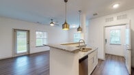 This single-floor residence offers a thoughtfully designed open-concept layout that seamlessly connects the kitchen and living areas. The kitchen serves as the centerpiece of the space, anchored by a peninsula with granite countertops that provide generous prep and seating space. A double stainless steel sink sits beneath a pair of brushed gold pendant lights, adding a warm, contemporary touch. White shaker-style cabinetry, a subway tile backsplash, and a full suite of stainless steel appliances — including a range, microwave, refrigerator, and dishwasher — complete this well-appointed cooking space.

The open living area flows naturally from the kitchen and features rich wood-look flooring throughout, crisp white walls, and a ceiling fan that enhances comfort and airflow. Multiple windows allow natural light to fill the space, creating a bright and inviting atmosphere with views of the surrounding greenery.

A dedicated laundry area accommodates a full-size washer and dryer, offering added convenience. Additional storage is provided through wire shelving units and a utility closet housing the water heater. The clean, neutral design palette throughout makes the space versatile and move-in ready.