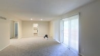 This single-floor space features a spacious, open-concept living area finished with soft, neutral-toned walls and plush wall-to-wall carpeting that adds warmth underfoot. Recessed lighting in the ceiling provides clean, even illumination throughout the room, complementing the natural light that pours in through a large sliding glass door fitted with vertical blinds. The sliding door offers direct access to an outdoor area, making it a functional and light-enhancing feature of the living space. A floor-level air vent is visible near the sliding door, indicating a built-in climate control system integrated into the flooring. The living area flows openly toward an adjoining room, visible through a doorway that leads to what appears to be a kitchen or utility area with cabinetry. The overall layout is straightforward and practical, with clean lines and minimal architectural detailing. The neutral color palette of beige walls and light gray carpeting creates a calm, versatile backdrop suited to a variety of furnishing styles. The space feels open and uncluttered, offering a functional and comfortable single-level living environment.