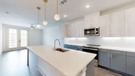 This well-appointed one-bedroom, one-bathroom residence is designed with a clean, modern aesthetic throughout. The open-concept kitchen and living area serves as the heart of the home, featuring a large center island with a white quartz countertop, an undermount sink, and stylish globe pendant lights suspended from brass fixtures. The kitchen is outfitted with two-tone cabinetry — light upper cabinets paired with slate-blue lower cabinets — accented by gold hardware, a classic white subway tile backsplash, and a full suite of stainless steel appliances including a side-by-side refrigerator, electric range, and microwave. Wood-look luxury vinyl plank flooring runs consistently throughout the unit, complementing the bright, neutral palette. The bedroom is a calm retreat with a ceiling fan, natural light from a window with blinds, and ample room to furnish comfortably. The walk-in closet features built-in shelving and hanging rods for well-organized storage. The bathroom impresses with a dual-sink vanity featuring grey cabinetry with gold pulls, a soaking tub, a separate glass-enclosed shower with tile surround, and access directly to the closet. A dedicated laundry room houses a full-size Whirlpool washer and dryer with overhead shelving.