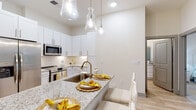 Located at 6146 Liberty Hall Dr in Leland, NC, this single-story residence offers a thoughtfully designed open floor plan with a clean, contemporary aesthetic. The kitchen serves as a central hub, showcasing crisp white shaker cabinetry, a classic subway tile backsplash, granite countertops, and a full suite of stainless steel appliances including a refrigerator with ice maker, microwave, and range. A large island with seating provides an ideal space for casual dining, illuminated by a trio of elegant glass pendant lights. Light wood-style flooring flows throughout the main living areas, complementing the bright, neutral color palette. The living area exudes a relaxed charm, with natural light filtering through floral-patterned drapes and ample wall space for art display. The primary bedroom is a serene retreat featuring soft carpet, warm neutral tones, and direct access to an en-suite bathroom. The bathroom is finished with wood-look tile flooring, a granite-topped vanity, and a tub-shower combination with tile surround. A generously sized walk-in closet with multi-tier wire shelving provides substantial storage. A second bedroom offers similar comfort, with carpeted floors, a bright window with views to greenery outside, and convenient access to a well-appointed full bathroom. The overall ambiance is light, airy, and move-in ready.