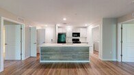This thoughtfully designed 2-bedroom, 2-bathroom residence in Harrisburg, PA offers a comfortable and modern living experience across a single floor. The open-concept kitchen and living area serves as the heart of the home, anchored by a large center island with quartz countertops, an undermount sink, and a contrasting wood-paneled base. The kitchen is outfitted with shaker-style cabinetry, a white subway tile backsplash, and black appliances including a gas range, microwave, and refrigerator. Wood-look plank flooring runs throughout the main living areas, lending warmth and continuity to the space. The living room is bright and airy, featuring a ceiling fan and sliding glass doors that open to a private balcony with outdoor views of surrounding greenery. Both bedrooms are carpeted and generously sized, each equipped with a ceiling fan. The spacious primary bedroom includes two closets, one of which is a walk-in with wire shelving. The two bathrooms are finished with light wood-tone flooring, gray shaker vanities, and illuminated mirrors. Additional storage is provided by two dedicated closets. The overall aesthetic is clean and contemporary, with a neutral color palette throughout.