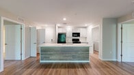 This thoughtfully designed 2-bedroom, 2-bathroom residence in Harrisburg, PA offers a comfortable and modern living experience across a single floor. The open-concept kitchen and living area serves as the heart of the home, anchored by a large center island with quartz countertops, an undermount sink, and a contrasting wood-paneled base. The kitchen is outfitted with shaker-style cabinetry, a white subway tile backsplash, and black appliances including a gas range, microwave, and refrigerator. Wood-look plank flooring runs throughout the main living areas, lending warmth and continuity to the space. The living room is bright and airy, featuring a ceiling fan and sliding glass doors that open to a private balcony with outdoor views of surrounding greenery. Both bedrooms are carpeted and generously sized, each equipped with a ceiling fan. The spacious primary bedroom includes two closets, one of which is a walk-in with wire shelving. The two bathrooms are finished with light wood-tone flooring, gray shaker vanities, and illuminated mirrors. Additional storage is provided by two dedicated closets. The overall aesthetic is clean and contemporary, with a neutral color palette throughout.