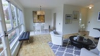This single-floor space offers a seamless open-plan layout that connects the living and dining areas, creating a generous and airy atmosphere throughout. The flooring is a standout feature — classic parquet tiles in a warm honey tone run continuously across the space, lending it a timeless and refined character.

The living area is anchored by a geometric patterned area rug in soft grey and white, complemented by a sleek white armchair and a dark oval coffee table. A tufted black leather daybed adds a contemporary accent near the window, where natural light filters in generously, brightening the entire space.

The dining area is defined by a white rectangular table surrounded by matching white chairs, positioned beneath an elegant multi-arm chandelier with a fabric shade. A large abstract artwork on the wall behind the dining set adds a creative focal point to this zone.

The walls throughout are finished in a clean, neutral light grey, providing a calm and versatile backdrop. Framed artworks are thoughtfully placed along the walls, contributing to an understated yet curated aesthetic. A white interior door leads to an adjoining room, suggesting additional private spaces beyond the open living area. Overall, the space combines modern design sensibility with a warm and inviting ambiance.