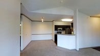 This single-floor residence offers a clean and functional open-concept design that seamlessly connects the living area to the kitchen. The living space is finished with plush wall-to-wall carpeting and features vaulted ceiling details accented with crown molding and a decorative border rail along the walls, adding a touch of architectural character. Track lighting illuminates the space, complementing the bright, neutral color palette throughout.

The kitchen is well-appointed with rich dark cabinetry, a stainless steel microwave, a range, and a full-size refrigerator with a freezer. A peninsula-style breakfast bar with a granite-look countertop serves as a natural divider between the kitchen and the living area, offering additional counter space and casual seating opportunities. The kitchen flooring transitions to a hard surface, providing a practical contrast to the carpeted living zone.

Overall, the space presents a comfortable and cohesive interior with a straightforward layout that maximizes usability. The vaulted ceiling detail in the living area enhances the sense of openness, while the integrated kitchen design makes it ideal for everyday living and casual entertaining.
