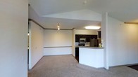 This single-floor residence offers a clean and functional open-concept design that seamlessly connects the living area to the kitchen. The living space is finished with plush wall-to-wall carpeting and features vaulted ceiling details accented with crown molding and a decorative border rail along the walls, adding a touch of architectural character. Track lighting illuminates the space, complementing the bright, neutral color palette throughout.

The kitchen is well-appointed with rich dark cabinetry, a stainless steel microwave, a range, and a full-size refrigerator with a freezer. A peninsula-style breakfast bar with a granite-look countertop serves as a natural divider between the kitchen and the living area, offering additional counter space and casual seating opportunities. The kitchen flooring transitions to a hard surface, providing a practical contrast to the carpeted living zone.

Overall, the space presents a comfortable and cohesive interior with a straightforward layout that maximizes usability. The vaulted ceiling detail in the living area enhances the sense of openness, while the integrated kitchen design makes it ideal for everyday living and casual entertaining.
