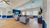Located at 401 W Seminole Blvd in Sanford, Florida, this single-story community space is thoughtfully designed with a cohesive coastal-inspired color palette, featuring bold navy blue wainscoting paired with soft sage-gray upper walls throughout. Light hardwood flooring runs continuously across the open layout, enhancing the bright and welcoming atmosphere.

The main lounge area is generously sized, furnished with multiple gray upholstered sofas and armchairs arranged around patterned area rugs. Ceiling fans and recessed lighting provide comfort, while an abundance of windows fitted with white plantation blinds allow natural light to fill the space. A wall-mounted flat-screen television serves as a central focal point in this gathering area.

An adjacent dining nook features a round glass-top table with seating for several guests and direct access to an exterior door. The compact kitchen is outfitted with white cabinetry, a stainless steel refrigerator, microwave, and countertop appliances, accented by a striking deep blue feature wall.

A dedicated private office is equipped with a large executive desk, multiple workstations, and ample window views of the surrounding greenery, making it well-suited for leasing or administrative functions. Throughout, the space maintains a polished, professional, yet inviting ambiance.