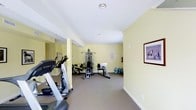 This fitness room offers a functional and well-organized space for exercise and wellness. The room is finished with soft yellow walls and white ceilings, creating a bright and energizing atmosphere. Neutral carpet flooring runs throughout, providing a comfortable surface underfoot.

The space is equipped with a range of exercise equipment, including a treadmill and elliptical machine positioned toward the front of the room for cardio training. A multi-station cable and weight machine occupies the central area, offering a variety of strength training options. Additional equipment, including a recumbent bike, is also available, catering to a range of fitness preferences.

Large wall mirrors are installed along one side of the room, enhancing the sense of space while allowing users to monitor their form during workouts. Water jugs are conveniently available near the wall, supporting hydration during exercise sessions. A fire extinguisher and posted notices reflect attention to safety standards. Framed artwork on the wall adds a subtle decorative touch, contributing to an inviting and well-maintained environment overall.