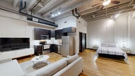 Located in the heart of downtown Dallas on Main Street, this urban loft offers a compelling blend of industrial character and modern comfort. The open-concept layout seamlessly connects the living, kitchen, and sleeping areas, all unified by warm hardwood flooring throughout. Soaring ceilings with exposed concrete beams, visible ductwork, and track lighting give the space a distinctive loft aesthetic that pays homage to its architectural roots.

The kitchen is well-appointed with dark espresso cabinetry, stainless steel appliances including a refrigerator, electric range, and microwave, complemented by a clean white countertop and undermount sink. The living area provides generous room for a sectional sofa and lounge furniture, anchored beneath the dramatic ceiling height.

The sleeping area occupies its own defined zone within the open floor plan, offering ample space for a full-size bed and nightstands, with a dedicated closet nearby. Natural light filters in through a framed window with bold dark trim, adding contrast to the bright white walls.

The bathroom features classic black-and-white penny tile flooring, a walk-in shower with white tile surround, and built-in open shelving for storage. A frosted glass door adds a stylish, private touch to the space. This loft delivers a distinctive urban living experience in one of Dallas's most vibrant neighborhoods.