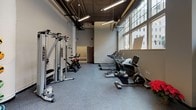 This single-floor fitness center is designed to support a comprehensive workout experience, combining thoughtful layout with a strong selection of equipment. The space features soaring ceilings with exposed ductwork and industrial-style pendant lighting, lending the room an open, energetic atmosphere. A generous bank of large, multi-pane windows lines one wall, flooding the interior with abundant natural light and offering views of the surrounding urban streetscape.

The flooring throughout consists of durable rubber gym matting, ideal for high-impact activities. Full-length mirrors span a significant portion of the walls, visually expanding the space and supporting form-conscious training. A wall-mounted flat-screen television is positioned for visibility across the workout floor.

Equipment is well-organized and varied. A dedicated free weights zone houses a full rack of dumbbells alongside kettlebells and an adjustable bench. A cable and pulley machine system supports strength training, while plyometric boxes of varying heights are available for functional fitness work. The cardio section lines another portion of the space and includes multiple treadmills, stationary bikes, and an elliptical trainer. Stability balls and resistance accessories round out the available equipment. The overall ambiance is clean, bright, and purpose-built for fitness.