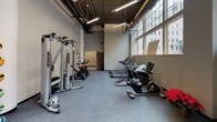 This single-floor fitness center is designed to support a comprehensive workout experience, combining thoughtful layout with a strong selection of equipment. The space features soaring ceilings with exposed ductwork and industrial-style pendant lighting, lending the room an open, energetic atmosphere. A generous bank of large, multi-pane windows lines one wall, flooding the interior with abundant natural light and offering views of the surrounding urban streetscape.

The flooring throughout consists of durable rubber gym matting, ideal for high-impact activities. Full-length mirrors span a significant portion of the walls, visually expanding the space and supporting form-conscious training. A wall-mounted flat-screen television is positioned for visibility across the workout floor.

Equipment is well-organized and varied. A dedicated free weights zone houses a full rack of dumbbells alongside kettlebells and an adjustable bench. A cable and pulley machine system supports strength training, while plyometric boxes of varying heights are available for functional fitness work. The cardio section lines another portion of the space and includes multiple treadmills, stationary bikes, and an elliptical trainer. Stability balls and resistance accessories round out the available equipment. The overall ambiance is clean, bright, and purpose-built for fitness.