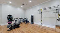 Located at 650 Dekalb St in Auburn, Alabama, this compact yet fully outfitted fitness studio offers a comprehensive workout environment within a single, open-plan space. The room features warm-toned hardwood flooring throughout, light gray walls with crisp white crown molding and baseboards, and recessed ceiling lighting that creates a clean, well-lit atmosphere conducive to exercise.

The studio is thoughtfully organized to support a variety of workout styles. A row of stationary spinning bikes lines one wall, ideal for cycling-based cardio training. A freestanding heavy punching bag provides options for boxing and striking workouts, while a wall-mounted horizontal bar rack with TRX suspension straps supports functional strength and bodyweight training. A wall-mounted equipment organizer holds medicine balls, resistance bands, and other accessories for added versatility.

On the free weights side, the studio is stocked with a tiered dumbbell rack and a double-tier kettlebell rack offering a range of weights. A large interior window looks into an adjacent room where treadmills are visible, and a glass-paneled door connects to additional areas of the facility. A wall-mounted mirror expands the sense of space and allows for form monitoring during workouts. Wall art and a digital thermostat round out the space, making this studio both functional and welcoming.