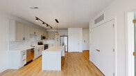 Located at 1116 Missouri Ave in St. Louis, MO, this thoughtfully designed single-floor unit offers a clean, modern aesthetic with an open-concept layout that seamlessly connects the kitchen and living areas. The kitchen is a standout feature, showcasing crisp white shaker-style cabinetry extending to the ceiling, a white subway tile backsplash, and a full suite of stainless steel appliances including a range, microwave, dishwasher, and refrigerator. A large center island with a bright white countertop provides ample workspace and serves as a natural focal point, illuminated by a stylish cluster of globe pendant lights suspended from a black track rail. Light-toned hardwood flooring runs throughout the main living area, lending warmth and cohesion to the space. The living area benefits from natural light streaming through windows that offer views of the surrounding neighborhood. The bedroom features plush carpeting and a generously sized walk-in closet with built-in shelving, offering practical storage solutions. The bathroom is finished in a clean, contemporary style with a white vanity, framed mirror, and matching cabinetry hardware consistent with the rest of the unit. Overall, the space presents a bright, airy ambiance with a consistent modern design language throughout.