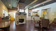 This single-floor space makes a strong first impression with its thoughtful blend of rustic charm and functional design. The centerpiece of the main area is a commanding floor-to-ceiling stone fireplace, complete with a solid wood mantel and an elegantly arched firebox opening. A flat-screen television is mounted directly above the mantel, making the fireplace wall both a focal point and a practical feature. Overhead, richly textured wood-beam ceilings add depth and a warm, lodge-inspired character to the space, complemented by recessed lighting that ensures the interior remains well-lit. The flooring throughout is a deep, dark-stained hardwood that anchors the room with a sense of solidity and refinement. A seating area with ornate wooden chairs and a small side table creates an inviting nook for relaxed conversations or waiting. Toward the back of the space, a built-in counter with overhead cabinetry and a mirror suggests a reception or service area, adding a professional and organized element to the layout. Framed artwork adorning the walls contributes to the curated, welcoming atmosphere. Overall, the space strikes a balance between rustic aesthetic and polished utility, making it equally suited for professional and social interaction.