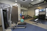 This fitness center offers a comprehensive workout environment designed to support a wide range of exercise routines. The space is organized into distinct functional zones, each tailored to a specific type of training. The cardio area features a row of treadmills and elliptical machines positioned near large windows with slatted blinds, allowing natural light to fill the room while offering outdoor views. Ceiling fans mounted within elegant tray ceilings help maintain a comfortable atmosphere throughout.

The strength training zone is well-appointed with a full dumbbell rack, adjustable benches, a power rack with weight plates, and mirrored walls that enhance the sense of openness. A standout feature is the vibrant orange functional training rig, equipped with resistance bands, stability balls, medicine balls, and kettlebells, making it ideal for dynamic and circuit-based workouts. Additional cable machines and selectorized equipment provide options for targeted muscle training.

The flooring transitions between rubberized sport surfaces and lighter composite flooring, clearly delineating workout zones while providing safety and durability. Recessed lighting combined with ceiling fans ensures the space remains bright and well-ventilated. Glass doors open to an outdoor balcony area, adding a refreshing connection to the exterior. Overall, the fitness center is modern, functional, and thoughtfully arranged to accommodate a variety of fitness goals.