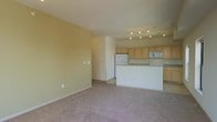 This single-floor residence features an open-concept design that seamlessly connects the living area to the kitchen. The living space is finished with wall-to-wall carpet in a neutral tone, complemented by warm yellow-beige walls that contribute to a bright and welcoming atmosphere. A low partial wall separates the living area from the kitchen without fully enclosing the space, maintaining an airy, connected feel throughout.

The kitchen is well-appointed with light wood cabinetry offering ample storage, a white refrigerator, a built-in microwave, and a range. A breakfast bar along the partial wall provides a casual dining or workspace option. Track lighting installed on the kitchen ceiling ensures the area is well-lit and functional. The kitchen flooring transitions from the carpeted living area to a hard surface, clearly delineating the two zones. Overall, the space presents a clean, practical layout suited for everyday living, with a consistent neutral color palette tying the rooms together cohesively.