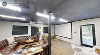 Located at 500 E Hargett St in downtown Raleigh, NC, this single-floor media and lounge space offers a well-appointed, multi-functional layout designed for both relaxation and productivity. The open floor plan is anchored by a large projection screen paired with a ceiling-mounted projector, making it ideal for presentations, screenings, or entertainment. A generous wraparound tufted brown leather sectional sofa dominates the central lounge area, complemented by a cowhide rug, a hairpin-leg coffee table, and additional green velvet seating — creating a comfortable and stylish gathering atmosphere. The dark, textured ceiling with exposed elements provides an industrial contrast to the bright white walls and warm light wood flooring throughout. A dedicated coffee bar area features white cabinetry with butcher block countertops and a refrigerator, while a long communal desk with bar-style seating offers a functional workspace. A round white dining table with modern white chairs serves as an additional gathering spot. Glass-paned double doors provide access to an adjacent area, and a window along one wall brings in natural light. The space blends industrial, modern, and cozy design elements into a cohesive, highly functional environment.