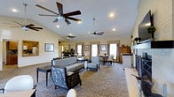Located at 680 Wildflower Drive in Wilkes-Barre, PA, this single-floor space offers an open and welcoming layout designed for comfort and functionality. The main gathering area features vaulted ceilings that enhance the sense of openness, complemented by multiple ceiling fans that provide both practical airflow and a finishing architectural touch. Recessed lighting is installed throughout, ensuring consistent illumination across the space.

The room is furnished with a thoughtful arrangement of seating, including a sofa, accent chairs, and side tables, creating multiple conversation areas suited for relaxed gatherings. Patterned upholstery on select furnishings adds visual interest against the neutral tones of the carpet and walls. Several windows along the perimeter allow natural light to filter in, brightening the interior while framed artwork on the walls adds a curated, polished feel.

Toward the rear of the space, a dining area with tables and chairs provides a designated spot for meals or meetings. A service counter or bar area is also visible, suggesting the space accommodates catering or refreshment needs. A wooden hutch or cabinet serves as a functional storage feature. Overall, the space presents a clean, well-appointed environment ideal for communal use.