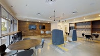 This contemporary single-floor communal space is thoughtfully designed to accommodate a variety of social and collaborative activities. The most prominent architectural feature is the full-height reclaimed wood accent wall, which anchors the room with a warm, rustic texture and serves as a backdrop for two wall-mounted flat-screen televisions — ideal for group viewing or presentations.

The space is organized into distinct seating zones that encourage both casual interaction and focused conversation. A round wooden table paired with modern molded chairs occupies one area, while a counter-height bar table with matching stools sits directly in front of the wood wall. A central seating cluster features high-back upholstered pod chairs in deep blue, offering semi-private lounge areas on a patterned area rug with yellow accents.

Overhead, a sleek multi-arm pendant light fixture serves as a design centerpiece, complemented by recessed ceiling lighting that provides even, bright illumination throughout. The polished concrete flooring adds an industrial edge that balances the warmth of the wood elements. In the background, a tiled accent wall hints at a kitchenette or breakroom area. The overall ambiance is modern, inviting, and versatile — well-suited for a residential amenity lounge or collaborative workspace.