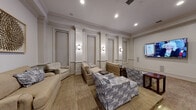 Located in Charlottesville, Virginia, this refined lounge space showcases classic architectural detailing throughout its single-floor layout. The room is defined by elegant raised-panel wainscoting that lines every wall, complemented by decorative pilasters and crisp crown molding that lend a formal, traditional character to the interior.

Recessed ceiling lighting works in harmony with a series of polished wall sconces, creating a warm, layered ambiance across the space. Neutral-toned carpeting anchors the room, while a wall-mounted flat-screen television serves as a central focal point on one end, supported by in-wall speaker panels that suggest a dedicated media experience.

Seating is arranged thoughtfully throughout, with a mix of plush sofas, armchairs, and accent chairs upholstered in coordinating beige and blue geometric-patterned fabrics. Small side tables and coffee tables in dark wood provide functional surfaces within each seating cluster.

An arched pass-through window on one wall adds architectural interest and hints at an adjacent service or reception area beyond. A framed sculptural wall accent provides a tasteful decorative touch, enhancing the overall refined aesthetic. The space exudes a welcoming, upscale atmosphere well suited for communal gathering and relaxation.