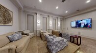 Located in Charlottesville, Virginia, this refined lounge space showcases classic architectural detailing throughout its single-floor layout. The room is defined by elegant raised-panel wainscoting that lines every wall, complemented by decorative pilasters and crisp crown molding that lend a formal, traditional character to the interior.

Recessed ceiling lighting works in harmony with a series of polished wall sconces, creating a warm, layered ambiance across the space. Neutral-toned carpeting anchors the room, while a wall-mounted flat-screen television serves as a central focal point on one end, supported by in-wall speaker panels that suggest a dedicated media experience.

Seating is arranged thoughtfully throughout, with a mix of plush sofas, armchairs, and accent chairs upholstered in coordinating beige and blue geometric-patterned fabrics. Small side tables and coffee tables in dark wood provide functional surfaces within each seating cluster.

An arched pass-through window on one wall adds architectural interest and hints at an adjacent service or reception area beyond. A framed sculptural wall accent provides a tasteful decorative touch, enhancing the overall refined aesthetic. The space exudes a welcoming, upscale atmosphere well suited for communal gathering and relaxation.