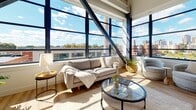 Located in Hartford, CT, this modern loft-style unit showcases an industrial-inspired design elevated by contemporary finishes. Soaring ceilings with exposed structural beams and ductwork set the tone, while dramatic floor-to-ceiling windows with black steel framing flood every room with natural light and frame sweeping views of the surrounding Hartford cityscape and skyline.

The open-concept living area is bright and airy, with light hardwood flooring that flows seamlessly throughout the space. The living room benefits from expansive corner windows and angled structural steel beams that add striking architectural character. The kitchen is a standout feature, boasting bold navy-blue cabinetry, a matching island with bar seating, light quartz countertops, stainless steel appliances, and pendant lighting — all framed by large windows overlooking the city.

The bedroom is generously proportioned, offering wall-to-wall window views and a serene, light-filled atmosphere. A dedicated home office area provides ample workspace, also bathed in natural light from oversized windows. The bathroom is clean and well-appointed, featuring large-format tile flooring, a wood-toned vanity with a white quartz countertop, and a full soaking tub. Throughout, the space balances industrial architectural elements with warm, modern finishes to create a refined urban living environment.