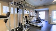 This fitness center offers a practical and well-appointed workout environment designed to support a range of exercise needs. The space is outfitted with a multi-station cable weight machine, providing versatile strength training options with an adjustable weight stack. A treadmill and an elliptical trainer offer dedicated cardio options, while a stationary exercise bike rounds out the cardiovascular equipment selection. The room features durable dark rubber flooring, ideal for both equipment placement and safety during workouts. Neutral-toned walls are accented by crown molding, giving the space a clean, finished appearance. Recessed lighting in the ceiling is complemented by ample natural light streaming through multiple windows fitted with blinds, creating a bright and energizing atmosphere. A wall-mounted flat-screen television is positioned for convenient viewing during exercise. Motivational fitness posters are displayed on the walls, adding an encouraging touch to the environment. The single-floor layout ensures easy accessibility, and the thoughtful arrangement of equipment maximizes the use of the available space without feeling cluttered. Overall, this fitness center provides a comfortable and fully functional setting for a comprehensive workout routine.