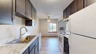 This single-floor space offers a practical and functional layout designed to maximize comfort and usability. The galley-style kitchen is a standout feature, outfitted with white shaker cabinetry extending to the ceiling for generous storage, granite-look countertops, and a full suite of black appliances including an electric range, over-the-range microwave, refrigerator, and dishwasher. A stainless steel sink with a modern faucet anchors the countertop workspace, while durable hardwood-style flooring runs the length of the kitchen, adding warmth and easy maintenance.

The living area is bright and open, with a large window that invites natural light and provides views of the surrounding neighborhood. Hardwood-style flooring continues here, complemented by baseboard heating and a wall-mounted air conditioning unit for year-round comfort.

The bedrooms are finished with plush carpeting and feature windows that bring in additional natural light. Sliding and hinged closet doors provide practical storage solutions, while baseboard heaters ensure consistent warmth. One bedroom offers convenient access to the bathroom, enhancing functionality.

Throughout the space, neutral wall tones create a clean, versatile backdrop, and the consistent design language across rooms lends a cohesive, move-in-ready feel.
