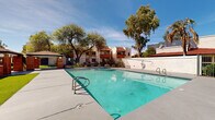 This two-story residential community is designed around a well-appointed outdoor common area that serves as its central gathering space. The architecture reflects a Southwestern aesthetic, characterized by stucco exteriors, red tile roofing, and warm earth tones that complement the surrounding desert landscape.

At the heart of the community is a generously sized swimming pool with crystal-clear turquoise water, framed by a spacious concrete pool deck ideal for lounging. An adjacent spa provides an additional relaxation option. The pool area is secured by wrought iron fencing, offering both safety and an open, airy feel.

A standout feature of the outdoor space is the modern outdoor kitchen and bar area, clad in warm wood paneling and equipped with a built-in grill. Nearby, two stylish pavilion structures with hipped roofs and globe lighting provide shaded gathering spots. Lush artificial turf lawns border portions of the amenity area, creating vibrant green accents year-round.

Mature eucalyptus, palm, and shade trees are distributed throughout the grounds, offering natural canopy and enhancing the resort-like ambiance. Manicured garden beds with desert-friendly plantings add color and texture to the landscaping. The two-story residential buildings surround the amenity core, with upper-level balconies overlooking the pool and greenery, creating a cohesive and inviting community environment under open desert skies.