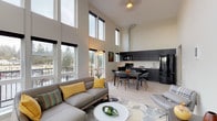 This contemporary loft-style residence makes an immediate impression with its dramatic double-height ceilings and expansive floor-to-ceiling windows that flood the open living area with natural light and offer sweeping views of the surrounding neighborhood. The light hardwood flooring flows seamlessly throughout the main living, dining, and kitchen areas, tying the open-concept layout together cohesively.

The living room is generously proportioned, accommodating a sectional sofa, lounge chair, and coffee table with room to spare, all anchored by a bold geometric area rug. The kitchen is outfitted with dark, floor-to-ceiling cabinetry, black appliances, and a clean-lined countertop with a single-basin sink, complemented by a dining area that seats four comfortably beneath the soaring ceiling.

The bedrooms continue the modern aesthetic with tall windows providing ample natural light and views. One bedroom features a striking loft space accessible via a metal ladder, adding a unique architectural element and extra functional space. Bathrooms are clean and functional, finished in neutral tones with chrome fixtures. A convenient in-unit stacked washer and dryer is tucked neatly within a dedicated closet. Exposed ductwork and wood-accent ceiling details add an industrial-chic character throughout the space.
