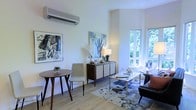 This single-floor space presents a thoughtfully designed interior that blends contemporary style with a warm, inviting ambiance. The main living area is bathed in natural light, courtesy of large windows that frame verdant outdoor greenery, creating a seamless connection between the interior and the natural surroundings outside.

The room features light hardwood-style flooring that enhances the bright, airy atmosphere throughout. A modern split-unit air conditioning system is mounted on the crisp white wall, ensuring year-round comfort. The living area is anchored by a patterned area rug and furnished with a sleek glass coffee table, an upholstered armchair, and a classic tufted accent chair, creating a balanced and functional seating arrangement.

A compact dining nook with a round wooden table and a minimalist chair offers an intimate space for casual meals. A mid-century modern sideboard adds both style and storage, topped with decorative accents and a soft table lamp. The walls are adorned with bold, expressive artwork that adds character and visual interest to the space.

Overall, the design aesthetic is clean and contemporary, with a careful mix of textures, tones, and artistic details that create a cohesive and welcoming environment throughout.