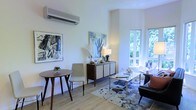This single-floor space presents a thoughtfully designed interior that blends contemporary style with a warm, inviting ambiance. The main living area is bathed in natural light, courtesy of large windows that frame verdant outdoor greenery, creating a seamless connection between the interior and the natural surroundings outside.

The room features light hardwood-style flooring that enhances the bright, airy atmosphere throughout. A modern split-unit air conditioning system is mounted on the crisp white wall, ensuring year-round comfort. The living area is anchored by a patterned area rug and furnished with a sleek glass coffee table, an upholstered armchair, and a classic tufted accent chair, creating a balanced and functional seating arrangement.

A compact dining nook with a round wooden table and a minimalist chair offers an intimate space for casual meals. A mid-century modern sideboard adds both style and storage, topped with decorative accents and a soft table lamp. The walls are adorned with bold, expressive artwork that adds character and visual interest to the space.

Overall, the design aesthetic is clean and contemporary, with a careful mix of textures, tones, and artistic details that create a cohesive and welcoming environment throughout.
