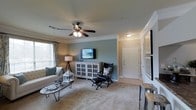 This single-floor space offers a comfortable and thoughtfully designed living environment. The living room serves as the central gathering area, showcasing elegant crown molding that frames the ceiling and adds a refined architectural touch. A ceiling fan with integrated lighting provides both comfort and ambiance, while plush wall-to-wall carpeting underfoot adds warmth to the space. The room is painted in a calming blue-grey accent tone, complemented by neutral walls that create a balanced and inviting atmosphere. Natural light filters in through a window dressed with patterned curtains, brightening the interior. A wall-mounted flat-screen television is positioned above a white, glass-paneled media cabinet, creating a clean and functional focal point. The seating area features a light-toned sofa and a classic leather chair with an ottoman, arranged around a sleek hairpin-leg coffee table. Near the entryway, a crisp white paneled door provides access to the space, flanked by a framed art display. The transition from carpeted living area to tile flooring at the entry adds a practical and visually distinct separation of zones. Overall, the space presents a harmonious blend of classic detailing and casual, livable comfort.